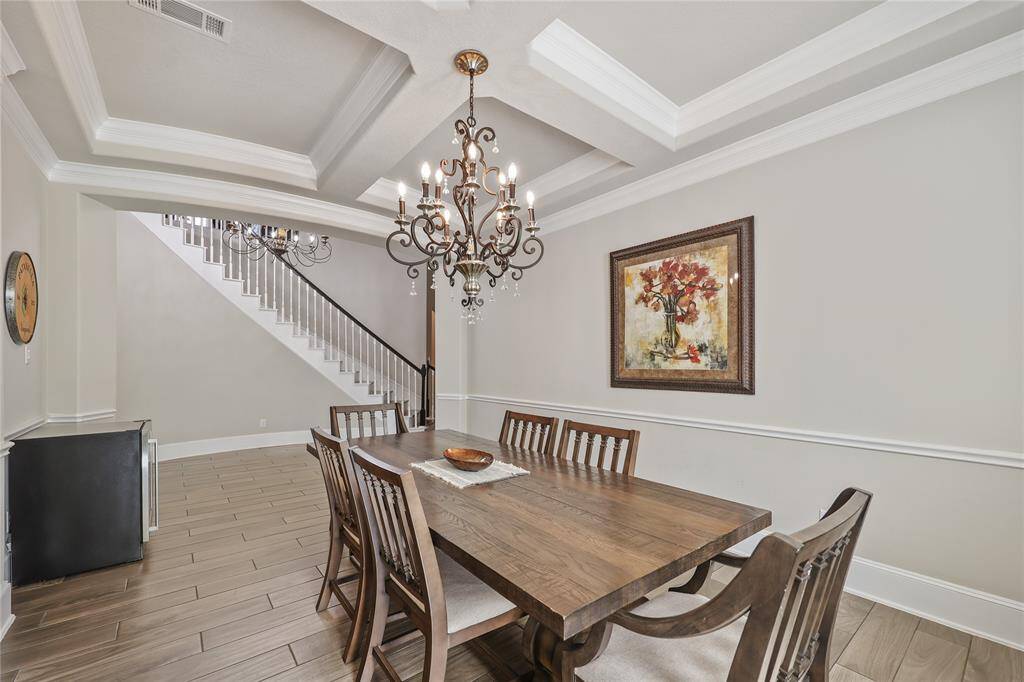 Experience elegance in this stunning formal dining room. A sophisticated chandelier with intricate detailing and crystal accents hangs from a coffered ceiling, adding timeless charm. The neutral-toned walls are complemented by elegant wainscoting paneling creating a warm and inviting ambiance.