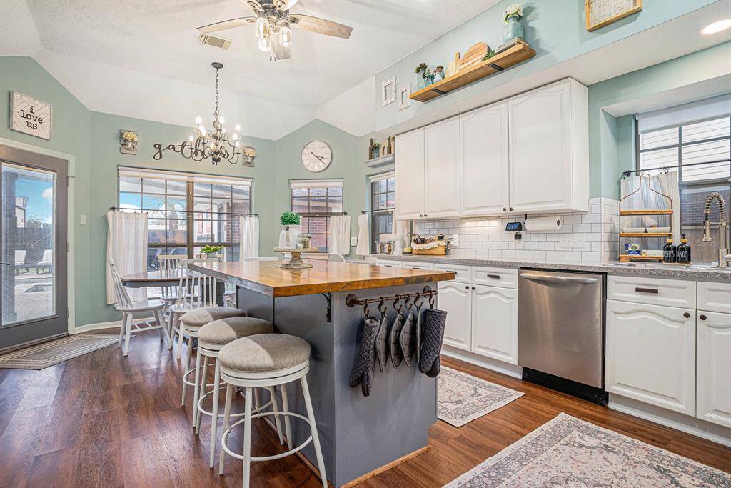 This kitchen features an inviting open layout with a central island, white cabinetry, and stainless steel appliances. The space is accented with warm wood floors, a chandelier, and ample natural light from large windows and breakfast area.