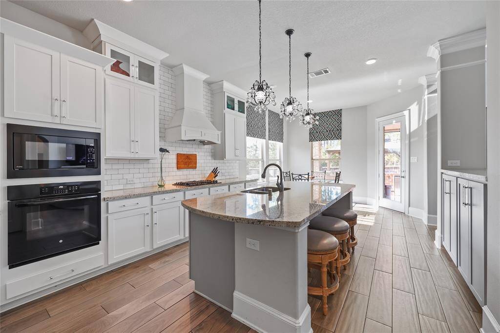 Beautiful gourmet kitchen featuring pristine white cabinetry, a sleek subway tile backsplash, and luxurious granite countertops. The high-end appliances, including a built-in microwave and oven, ensure culinary excellence. Sparkling pendant lighting adds a touch of sophistication, complementing the open-concept layout.  With warm wood-look flooring and ample natural light, this kitchen is the heart of the home!"