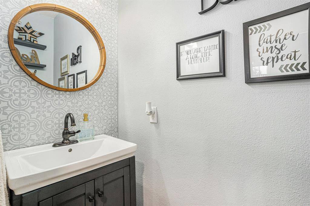 Half bathroom downstairs with stylish patterned wallpaper, round wooden-framed mirror, and a modern white sink on a black vanity.