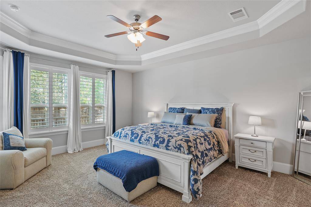 This primary bedroom features a spacious layout with a tray ceiling, adding depth and architectural interest to the room. A set of large windows with white plantation shutters allows ample natural light to enter, offering a bright and airy feel while maintaining privacy.