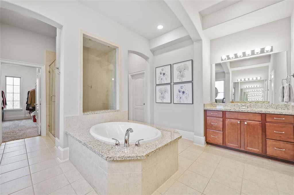 This primary bathroom showcases a luxurious soaking tub set into a granite surround, providing a spa-like ambiance. The large walk-in shower, enclosed by glass, features a built-in tiled niche for added convenience. Through the open doorway is one of two walk-in closets.