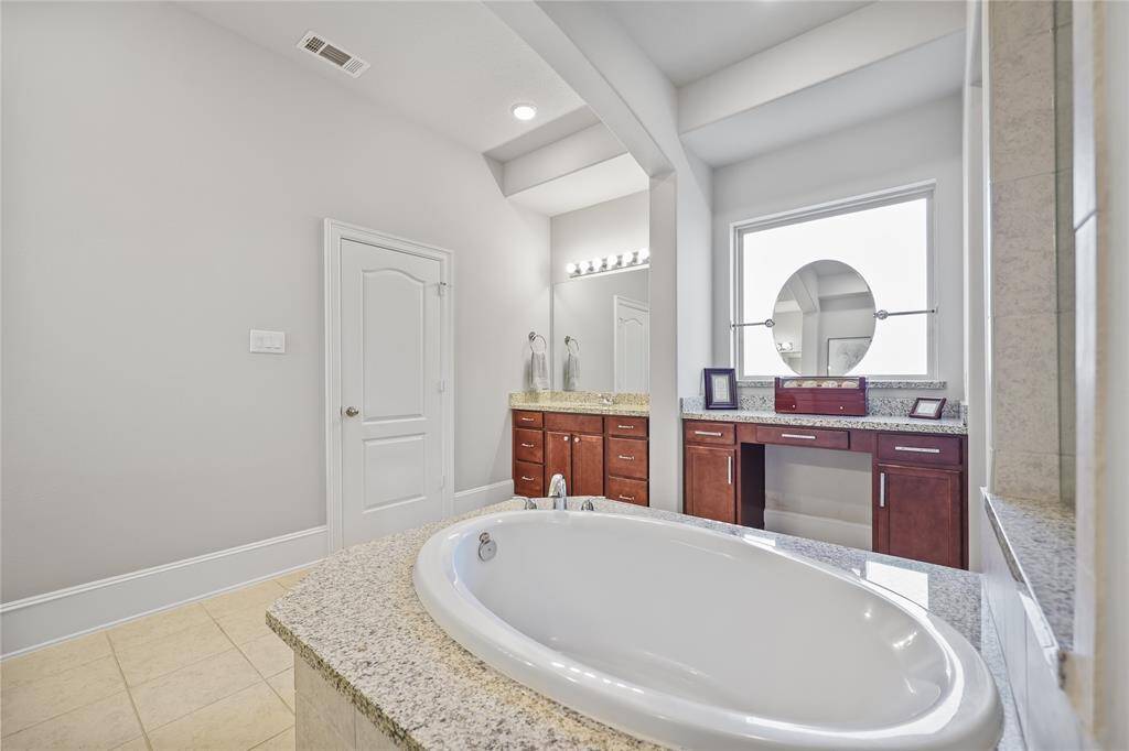 An elegant designed bathroom with a dual vanity setup, each with granite countertops and rich wood cabinetry, offering ample storage and counter space. A large framed mirror, illuminated by a row of bright vanity lights, enhances functionality and brightness. The flooring consists of large neutral-toned tiles, which seamlessly coordinate with the soft wall colors and granite surfaces.