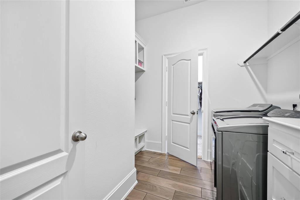 This utility room features wood-look tile flooring that provides durability and easy maintenance. The space includes built-in cabinetry and shelving, offering ample storage for laundry essentials. A long countertop with drawers/cabinets provides a practical workspace for folding and organizing, while overhead shelving with a hanging rod enhances storage options. The utility room has access from the entry through to one of the primary bedroom's walk-in closets.