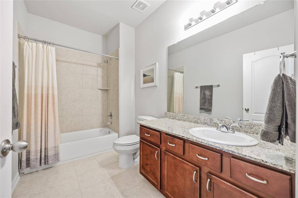 This upstairs hall bathroom features a granite countertop vanity and rich wood cabinetry for ample storage. A large mirror with bright vanity lighting enhances the space, creating a well-lit and open feel. The tub/shower combination is accented with neutral-toned tile surround, and the ceramic tile flooring complements the overall neutral color palette.