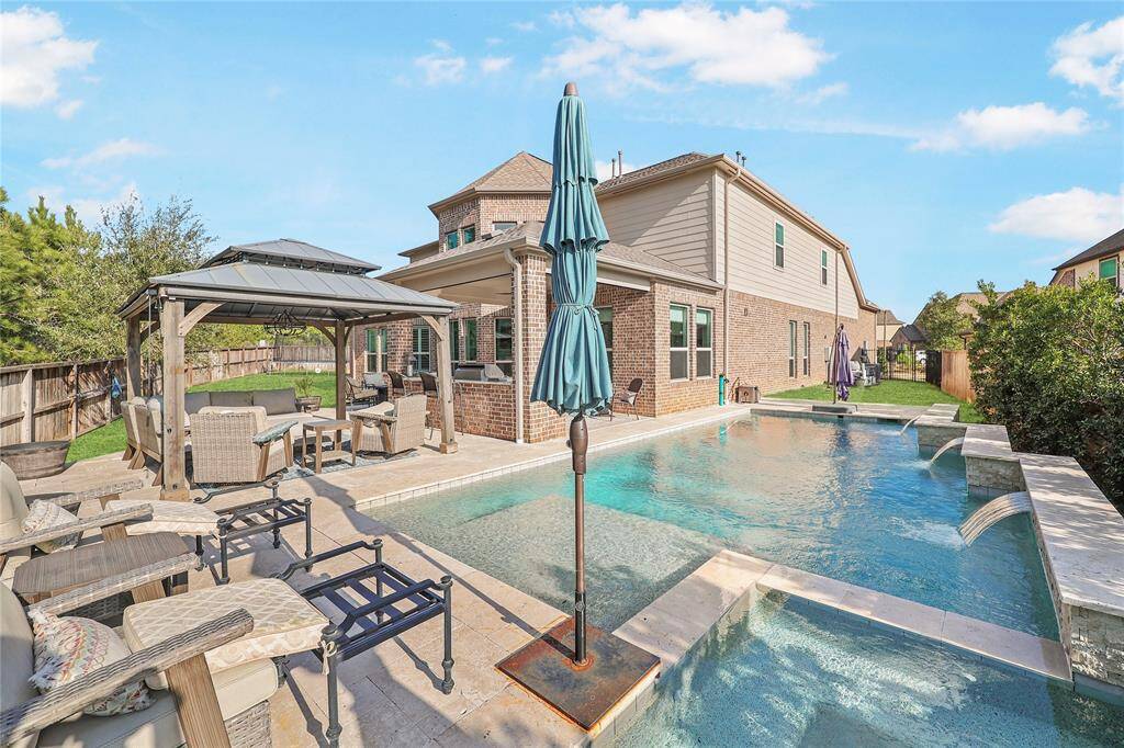 This luxurious backyard retreat features a resort-style pool with a built-in tanning ledge and elegant water features. The travertine tile pool deck extends seamlessly throughout the space, complementing the brick exterior of the home. A wooden pergola with a metal roof offers an additional outdoor lounging space, perfect for relaxation or social gatherings