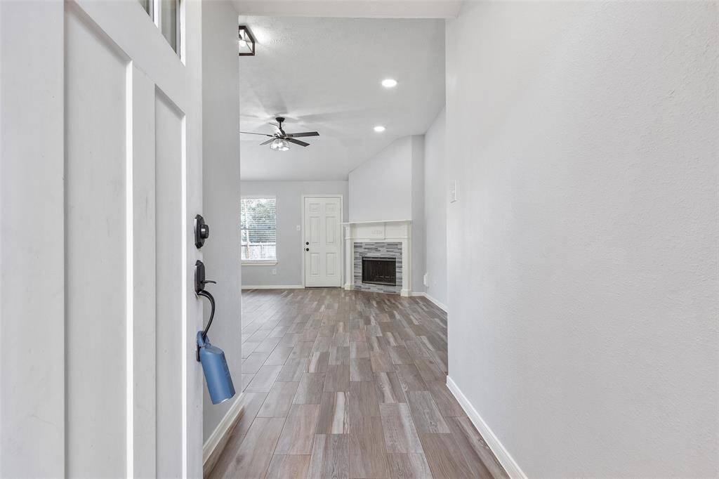 This photo shows a bright entryway leading into a spacious living area with wood-look tile flooring. The room features a modern ceiling fan, recessed lighting, and a cozy fireplace, creating a warm atmosphere.