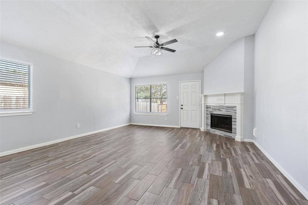 This spacious living room features stunning wood-look tile flooring, a cozy corner fireplace with a sleek design, and abundant natural light from large windows. The ceiling fan adds comfort and style to the room's airy atmosphere.