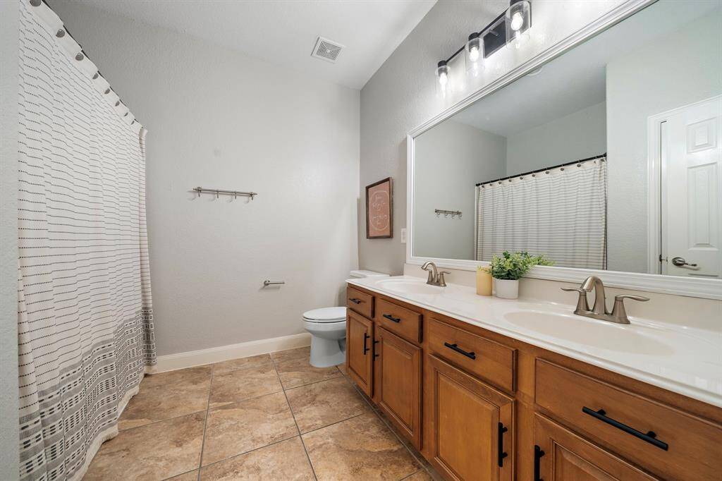 This spacious secondary bath combines functionality and style with a double sink, elegant vanity, and chic lighting. Tile flooring complements the design, offering a practical yet sophisticated space for daily routines.
