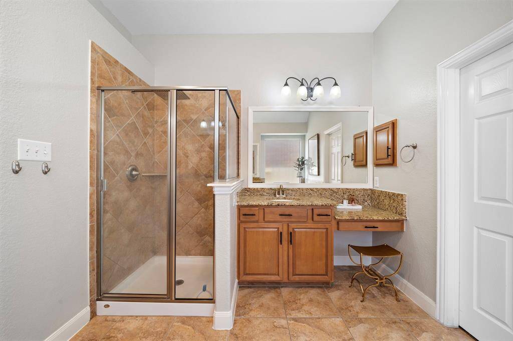 The ensuite bath offers a glass-enclosed shower with elegant tile work and dual granite vanities, blending functionality with luxury. Light wooden cabinetry provides ample storage, while a stylish vanity area adds a touch of elegance to this serene retreat.
