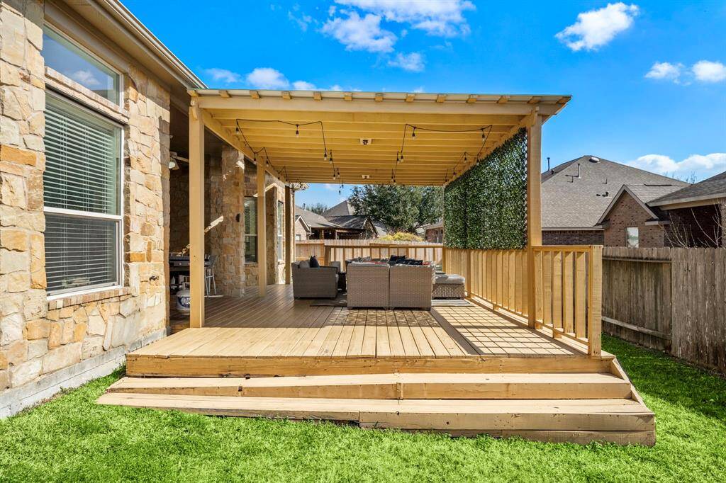 The spacious covered patio, enhanced with a pergola, offers a charming seating area for entertaining or relaxation. The wood decking complements the stone exterior and overlooks the lush green space, creating a seamless blend of indoor comfort and tranquil outdoor living.