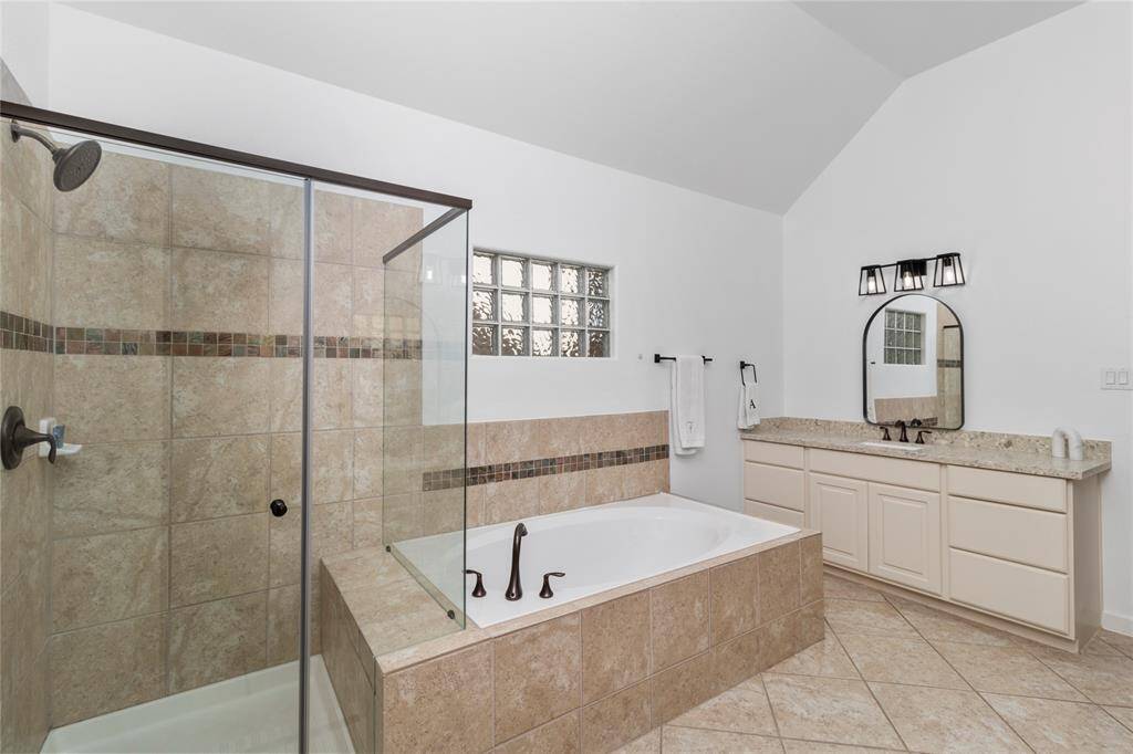This bathroom features a spacious walk-in shower with glass doors, a large soaking tub, and a dual-sink vanity with granite countertops. The neutral tilework and high ceiling create an open and elegant atmosphere.