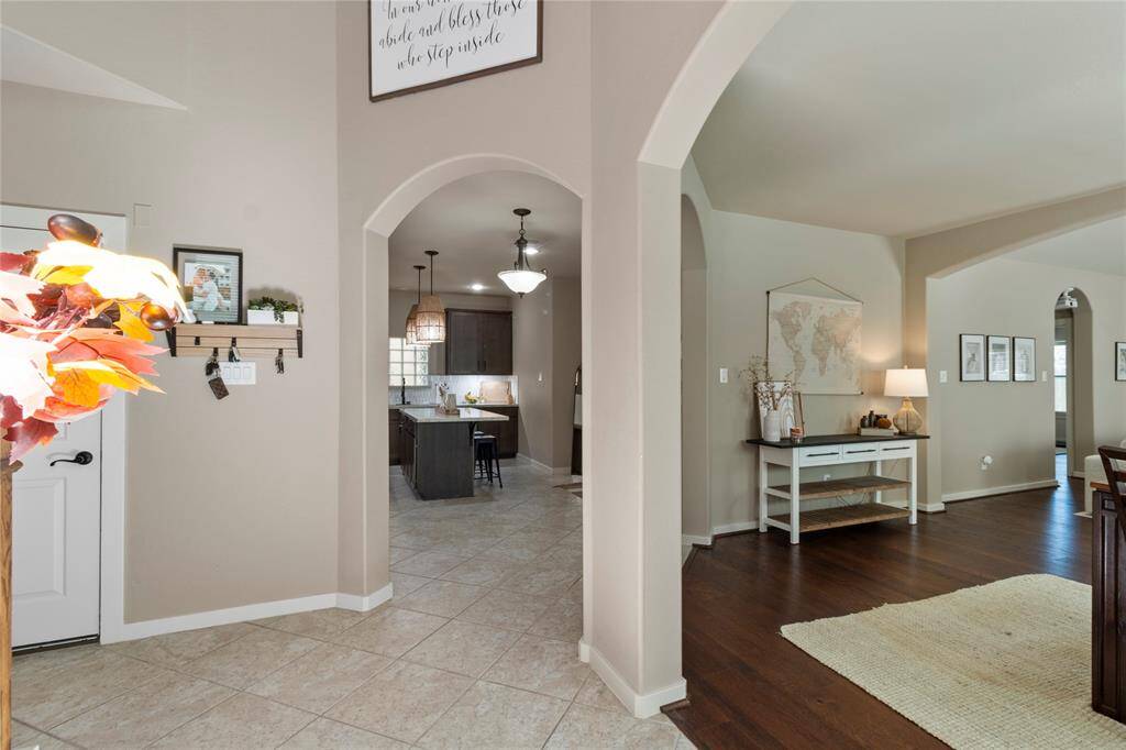 This photo showcases a welcoming entryway with neutral tones and tile flooring, leading into a cozy living area with hardwood floors. The kitchen features modern lighting and an open layout, visible through elegant archways. Stylish decor and ample natural light contribute to a warm and inviting atmosphere.