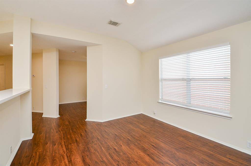 Bright room with hardwood floors, neutral walls, and a large window providing natural light. The open layout connects to another area, offering versatile living space.