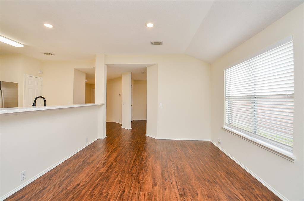 This photo showcases a bright, open room with wood laminate flooring and neutral walls. Large windows provide ample natural light, and the space features a modern, open layout with a view into an adjacent kitchen area.