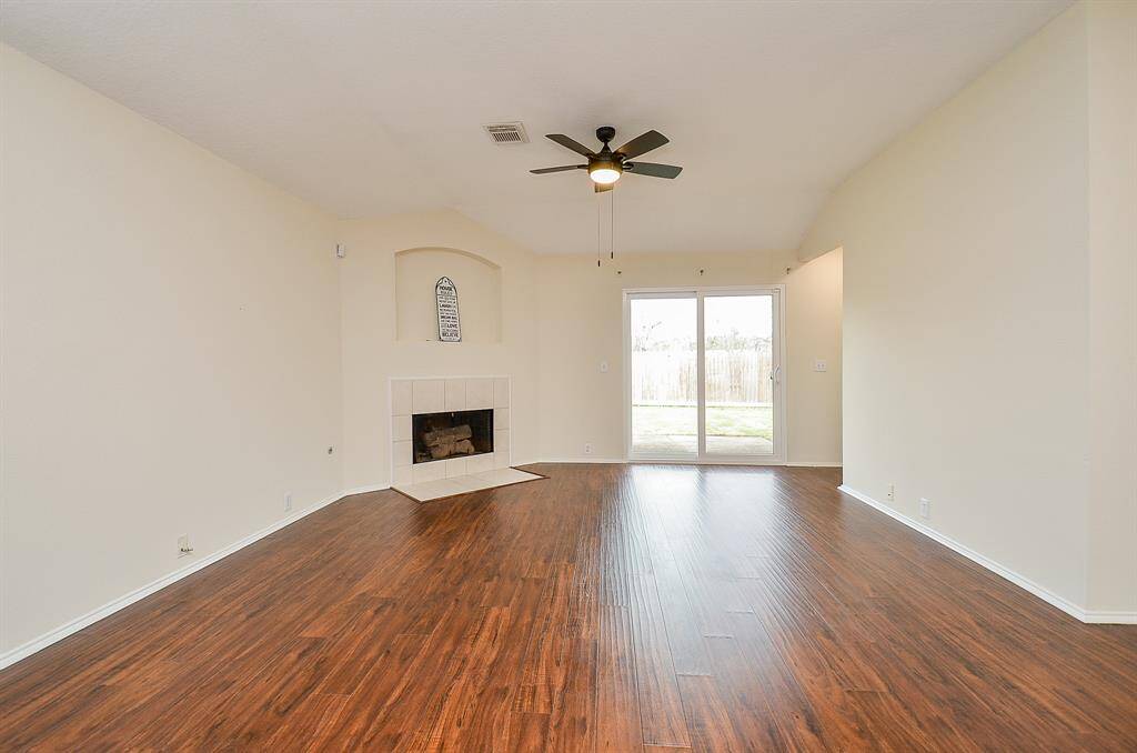 This spacious living room features warm wood flooring, a cozy fireplace, and ample natural light from large sliding glass doors leading to the backyard. A ceiling fan adds comfort and a modern touch to the bright, open space.
