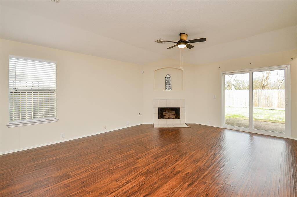 This spacious living room features elegant hardwood floors, a cozy white-tiled fireplace, and ample natural light from a large window and sliding glass doors leading to the backyard. The ceiling fan adds comfort to the airy, open space.