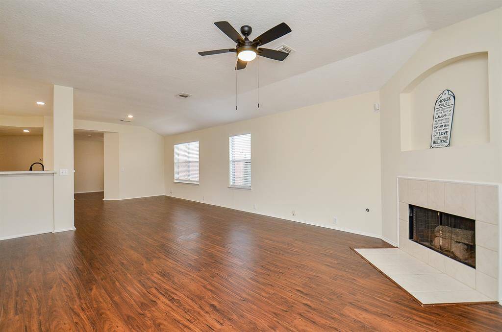 This spacious living room features sleek wood flooring, a cozy tile fireplace, and a ceiling fan. Natural light fills the room through two windows, and the open layout connects seamlessly to the adjacent kitchen area, creating a welcoming atmosphere.