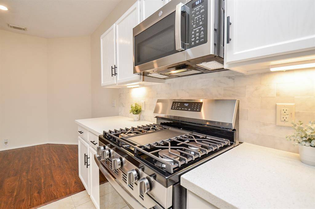 This kitchen features modern stainless steel appliances, including a gas stove and microwave. It has sleek white cabinetry, a light tile backsplash, and bright under-cabinet lighting, creating a clean and inviting cooking space.