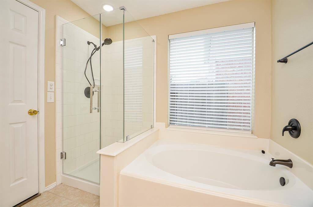 This bathroom features a sleek glass-enclosed shower and a spacious soaking tub, complemented by modern fixtures and a large window with blinds for natural light and privacy.