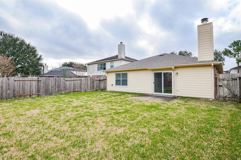 The photo shows a spacious backyard with a well-maintained lawn, enclosed by a wooden fence. The house features a light-colored exterior with a chimney and sliding glass doors leading to the patio. Ideal for outdoor activities and entertaining.