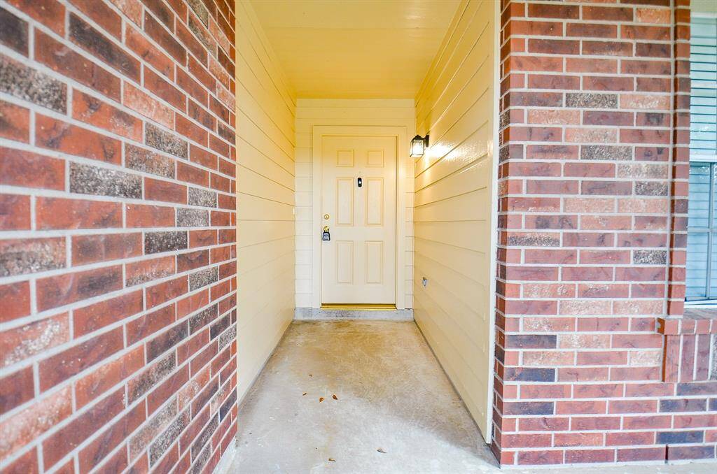This photo showcases a welcoming entryway with a covered porch, featuring a light-colored door and brick siding. A wall-mounted light fixture adds both style and security.