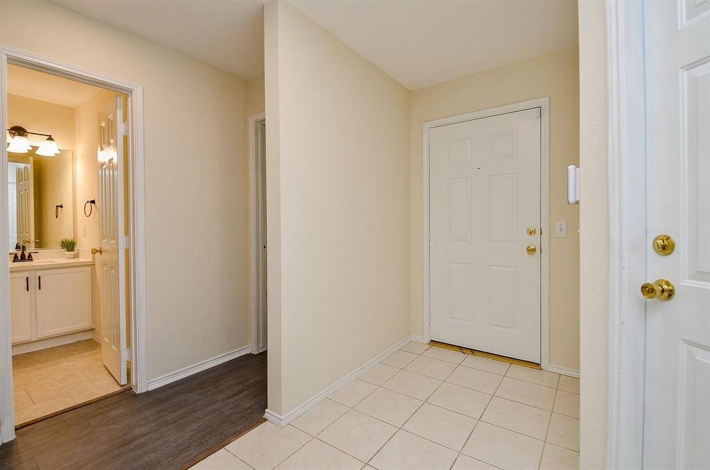 This photo shows a clean, welcoming entryway with neutral walls and tiled flooring. It leads to a bathroom with modern fixtures and a well-lit vanity. The space is well-maintained and inviting.