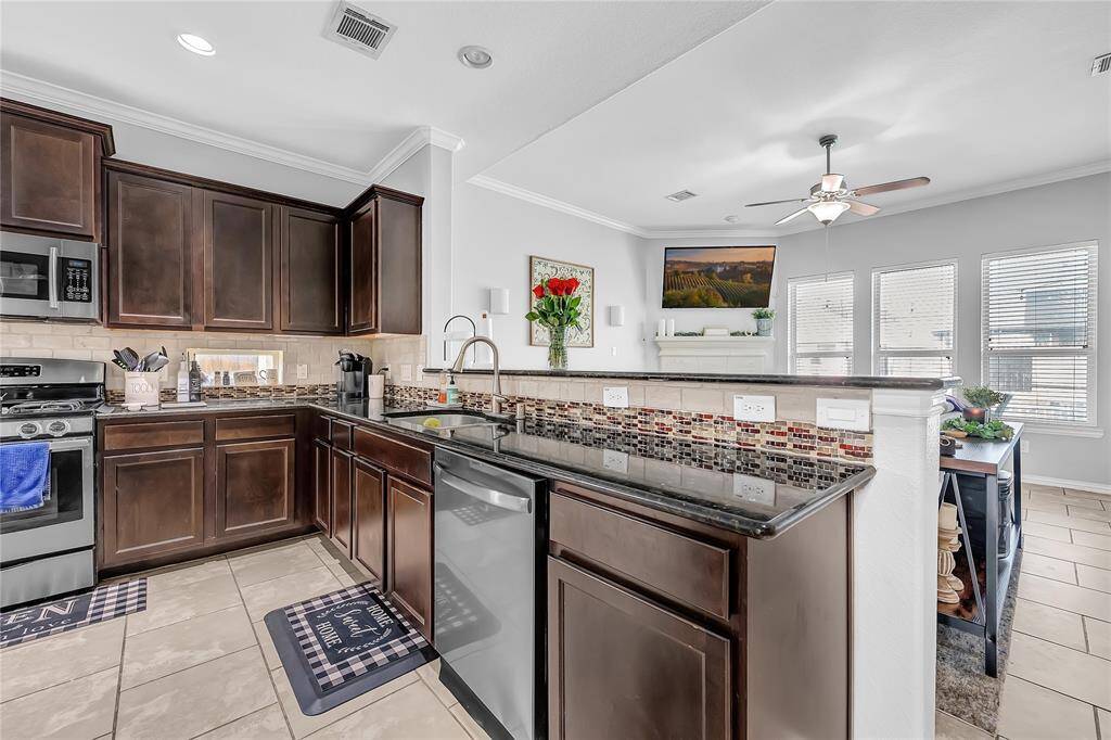 Modern kitchen with dark wood cabinets and stainless steel appliances. Features a stylish tiled backsplash and granite countertops. Open layout flows into a bright living area with large windows and ceiling fan.