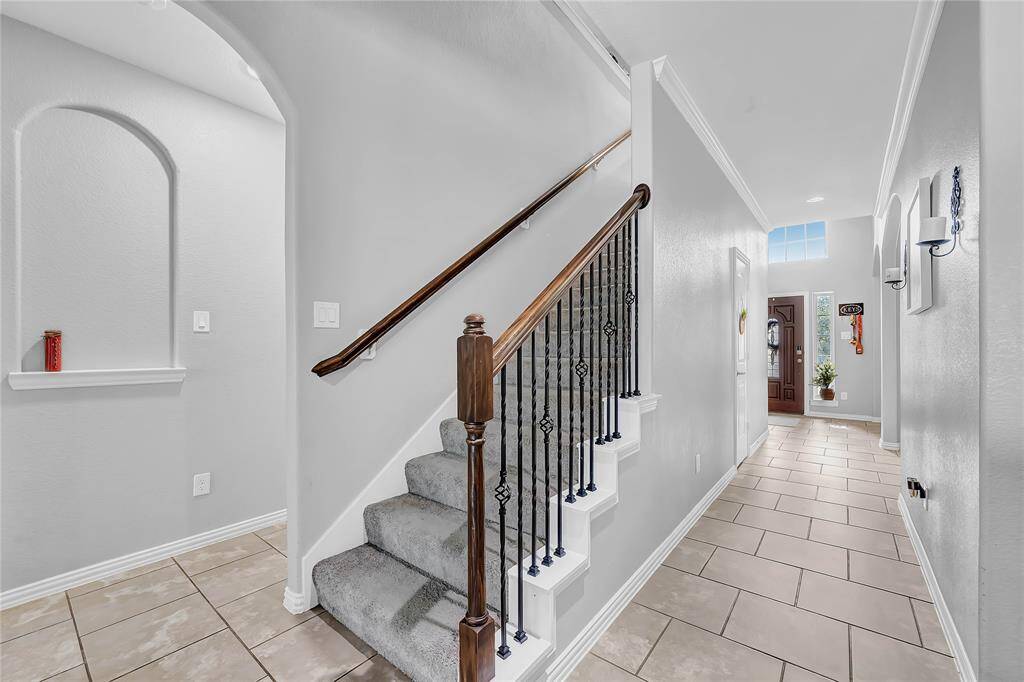 This inviting entryway features a bright hallway with tiled flooring, leading to a wooden staircase with wrought iron railings. The space is well-lit with natural light from the window near the front door, offering a warm and welcoming atmosphere.