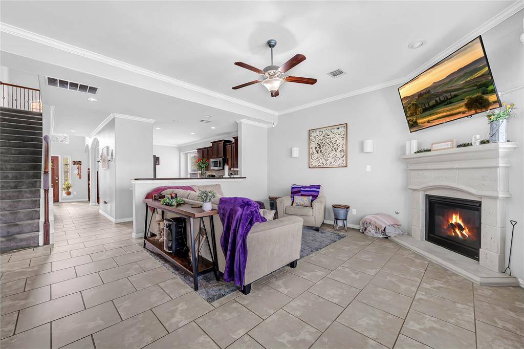 This spacious living area features a cozy fireplace, modern ceiling fan, and elegant tile flooring. The open floor plan seamlessly connects to the kitchen, while large windows provide ample natural light. The space is tastefully decorated with neutral tones and offers a comfortable, inviting atmosphere.