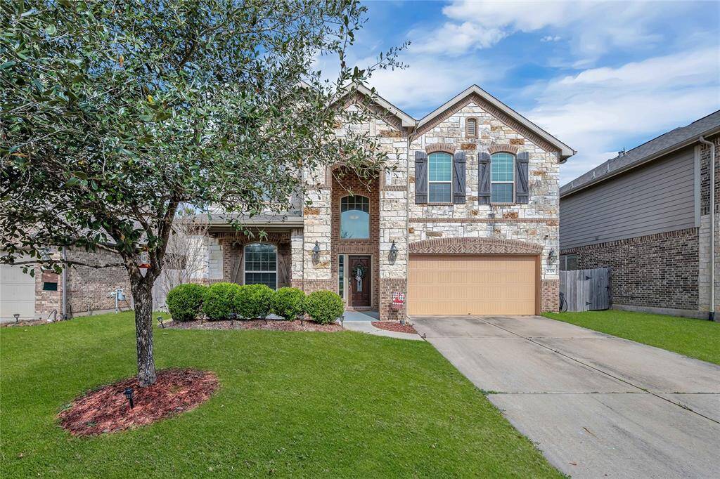 This is a two-story brick and stone house with a welcoming front entrance and a two-car garage. The well-maintained lawn features a mature tree and neat shrubs, creating an inviting curb appeal.