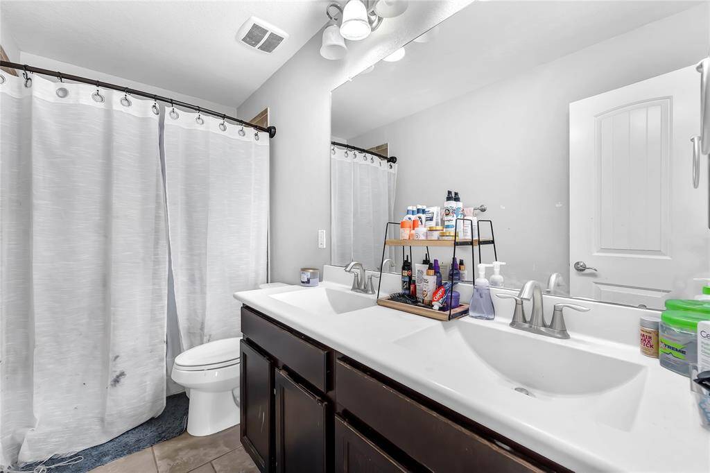 This bathroom features a double sink vanity with ample storage, a large mirror, and modern fixtures. It includes a shower with a curtain and neutral tile flooring, offering a clean and functional space.