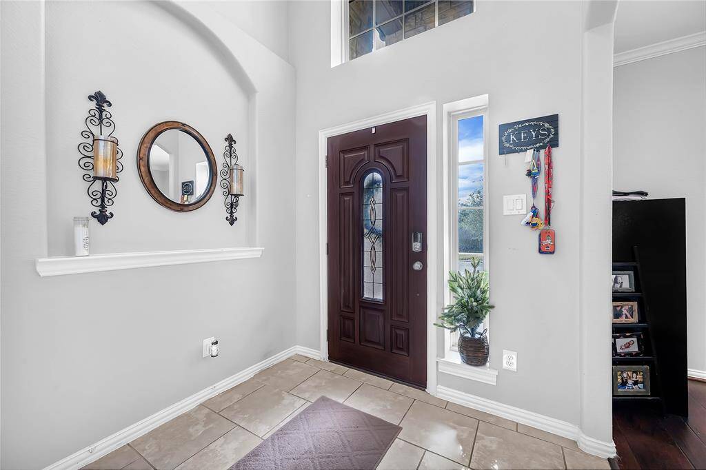 This welcoming entryway features a dark wood front door with decorative glass, flanked by windows for natural light. The neutral tile flooring and light gray walls create a bright atmosphere. Decorative wall accents and a small plant add charm, while a key holder provides functionality.