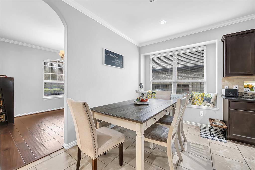 Bright dining area with a cozy window seat and modern decor. The space features a wooden table, cushioned chairs, and is adjacent to a sleek kitchen with dark cabinets. Tile flooring and ample natural light enhance the welcoming ambiance.