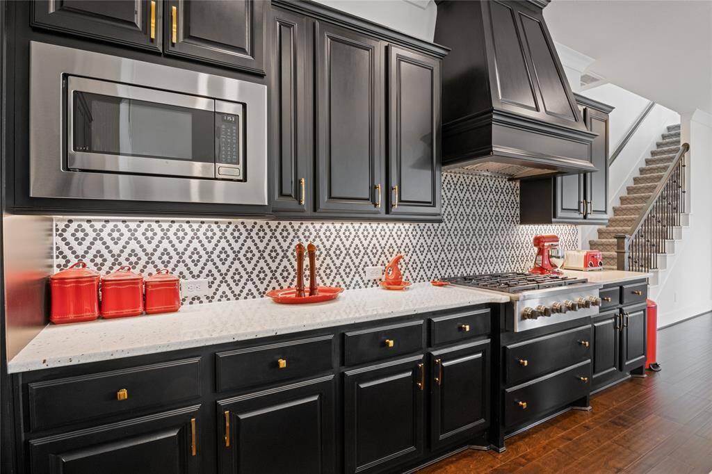 This kitchen features elegant black cabinetry with gold hardware and a sophisticated patterned backsplash. It includes modern stainless steel appliances, a spacious countertop, and stylish red accents, creating a vibrant and functional cooking space.