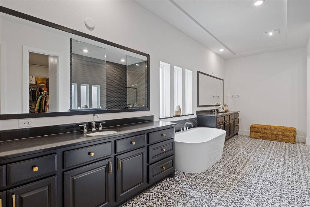 This elegant bathroom features a spacious layout with modern black cabinetry, a sleek freestanding tub, and decorative patterned tile flooring. It includes large mirrors, ample counter space, and natural light from multiple windows, creating a bright and luxurious atmosphere.