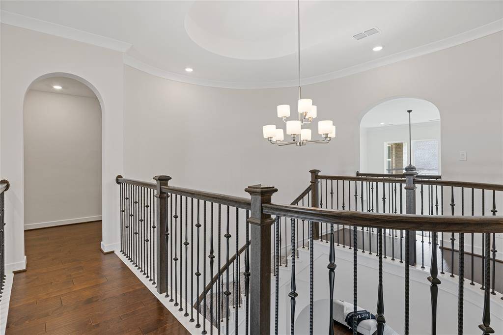 Stylish upper-level landing with rich wood flooring and elegant metal railings. The space features a modern chandelier, recessed lighting, and arched entryways, providing an open and inviting atmosphere.