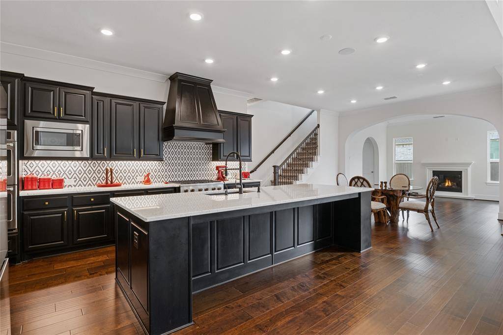 This home features an open-concept kitchen and living area with dark wood flooring. The kitchen boasts black cabinetry, a patterned backsplash, and a large island with a modern sink. The adjacent dining area leads to a cozy living space with a fireplace, creating an inviting atmosphere.