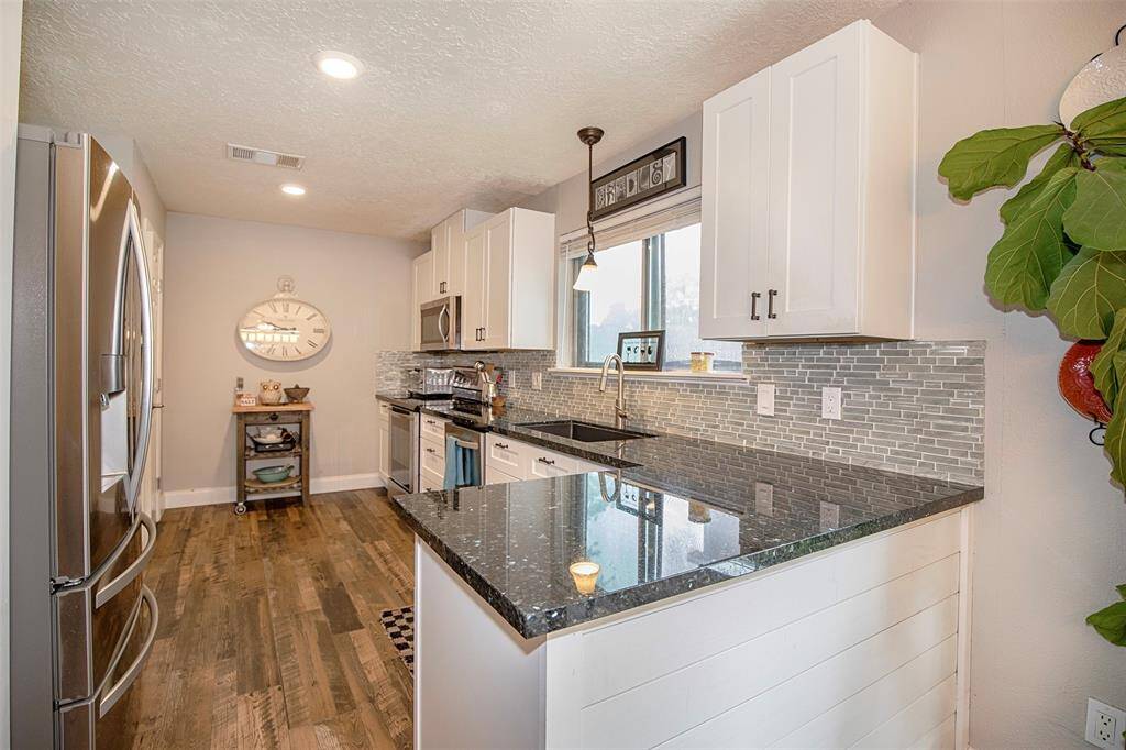 This photo showcases a bright dining area with a modern open-plan design, featuring an elegant lighting fixture and large window allowing natural light. The kitchen in the background is equipped with white cabinetry and stainless steel appliances, complemented by dark wood flooring.