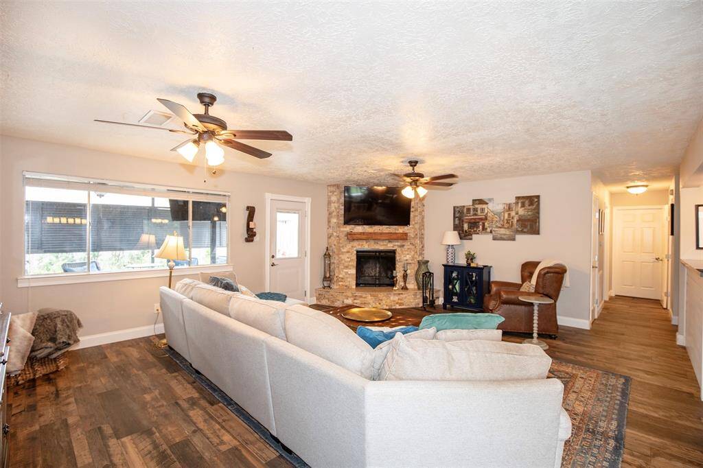This inviting living room features a cozy stone fireplace and warm wood flooring. It has a bright, open layout with ceiling fans and ample natural light from a wide window, creating a comfortable and stylish space.
