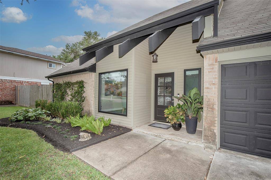 This photo showcases a modern home exterior with a clean, welcoming entrance. It features a covered entryway, a contemporary front door with glass panels, and a well-maintained garden with lush greenery. The house has a combination of brick and siding, complemented by a dark garage door.