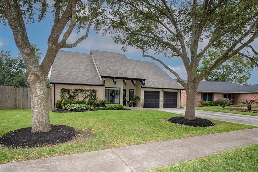 This photo shows a charming two-story home with a steep roof and brick exterior. It features a double garage, well-maintained lawn, and mature trees framing the entrance, creating an inviting curb appeal.