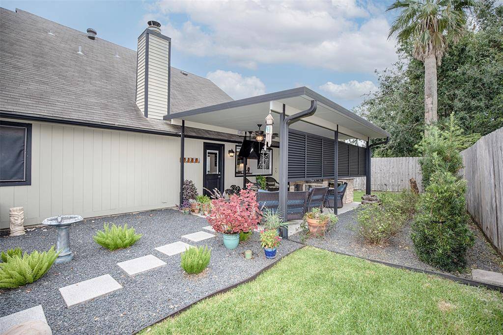 This photo showcases a cozy backyard patio with a covered seating area, surrounded by a well-maintained garden. The space features a modern pergola, lush greenery, and stepping stones leading to the house, creating a welcoming outdoor retreat.