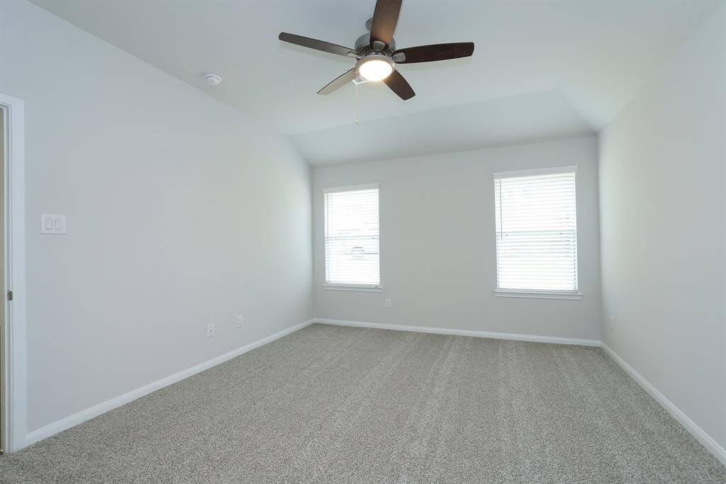 The soft gray walls,  plush carpet, and ample natural light from two windows with blinds creates a lovely sanctuary for the Primary Bedroom. A ceiling fan is centrally located, providing a modern touch and functional airflow.