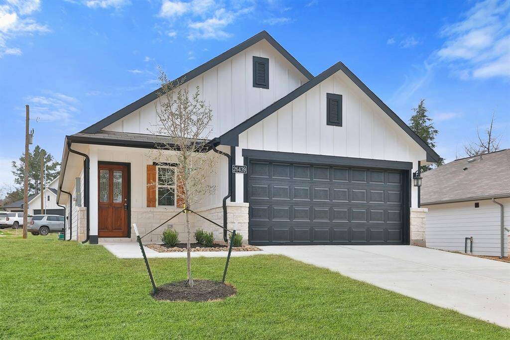 The clean, white exterior contrasts with black trim. It includes a two-car garage, a welcoming front porch with a wooden door, and a neatly landscaped yard with a newly planted tree. Ideal for contemporary living.