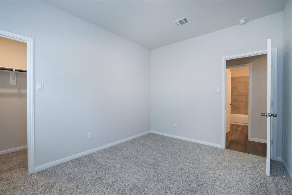 This is a bright, carpeted bedroom with neutral walls. It features a closet with a white door and an open doorway leading to a bathroom with vinyl plank flooring and modern lighting.
