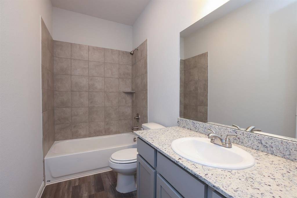 This bathroom features a modern design with a sleek granite countertop, a large mirror, and ample cabinet space. It includes a shower-tub combination, stylish tile surround and wood-look flooring, offering a clean and contemporary feel.
