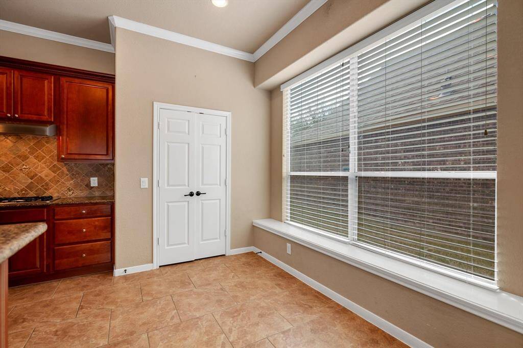 This photo shows a bright kitchen area with tan tile flooring and a large window with blinds, providing ample natural light. The kitchen features warm wooden cabinets, a tiled backsplash, and a white double door pantry.