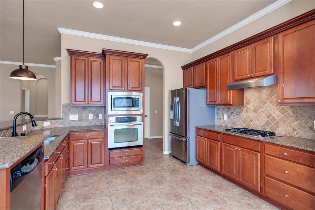This kitchen features elegant wood cabinetry, stainless steel appliances, and a stylish tile backsplash. It has ample counter space, a built-in oven, and a modern refrigerator, complemented by recessed lighting and a warm, inviting color scheme.