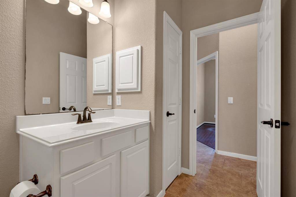 This photo shows a clean and modern bathroom with a white vanity and sink, bronze fixtures, and a large mirror. The space features neutral-colored walls and tile flooring, with a door leading to a hallway.